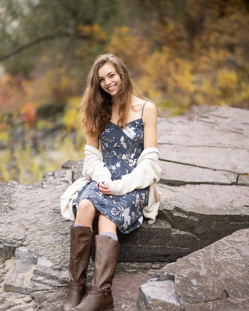 Alberton Senior Liberty Notley sits on a large rock with brown boots, a blue floral dress, and a white shawl. She has long brown hair and is smiling.